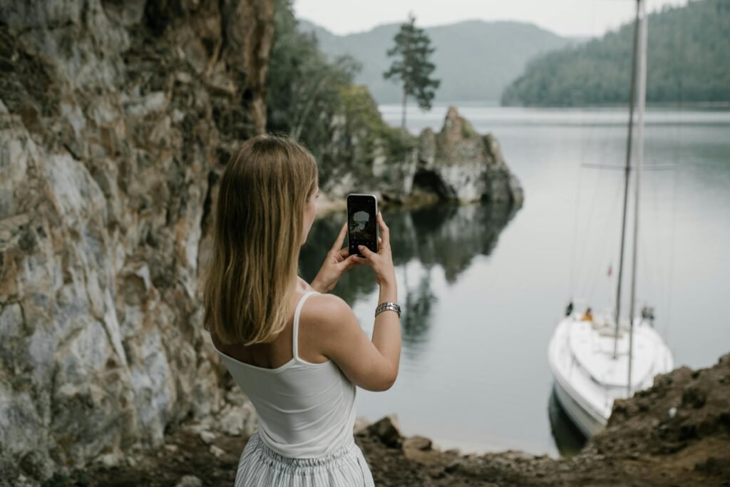 Viaje de autor: viaje diseñado con criterio y cuidado A woman takes a photo of a serene lakeside with a sailboat in lush natural surroundings.
