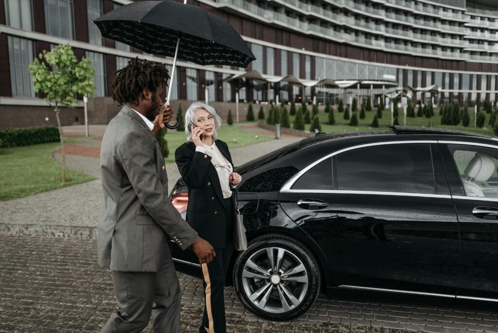 Two business professionals with a luxury car outdoors, using an umbrella and smartphone.